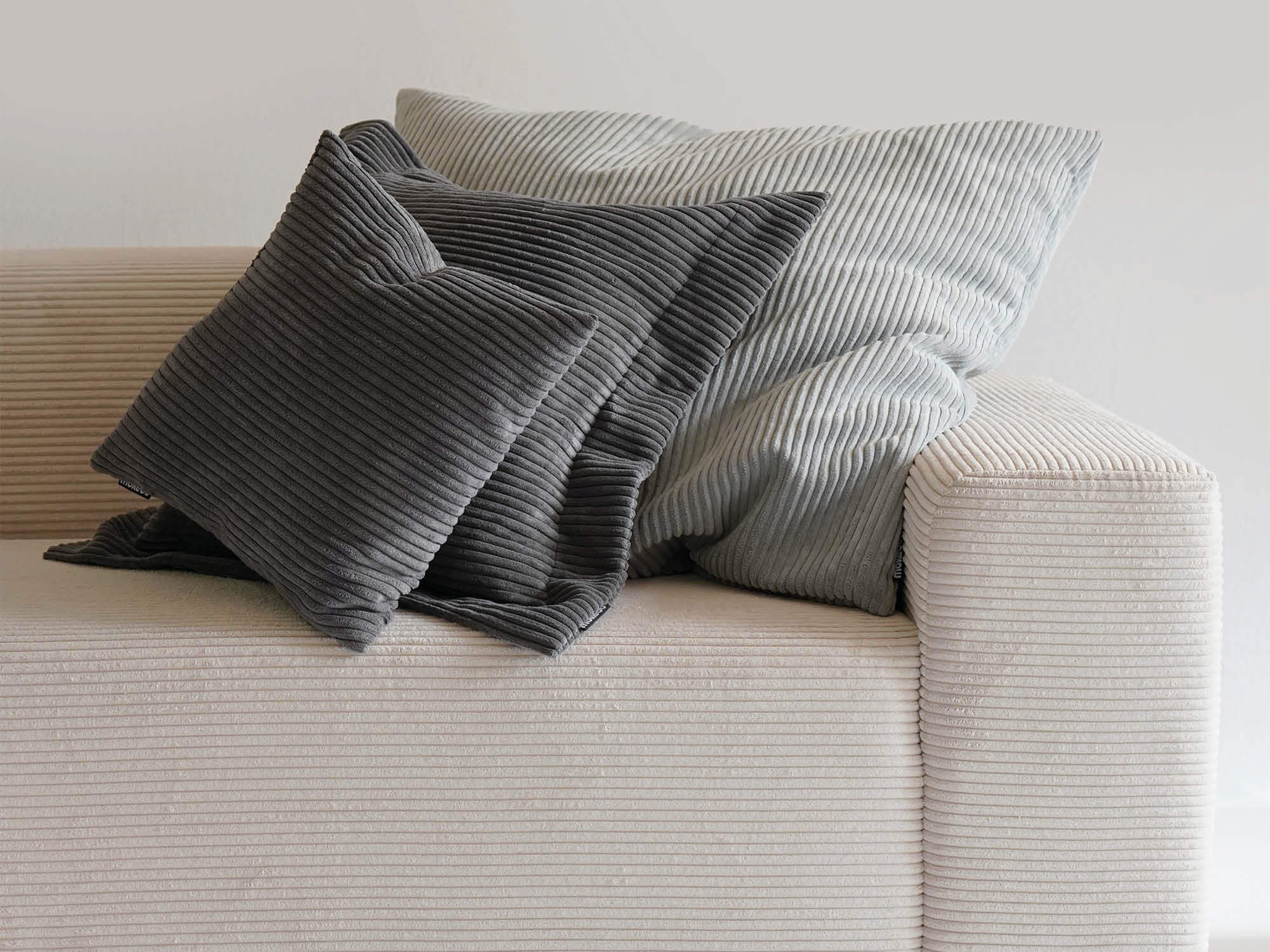 Drei unterschiedlich große Cordkissen hintereinander gelegt auf einem beigen Cordsofa. zwei sind anthrazit und ein Kissen ist grau