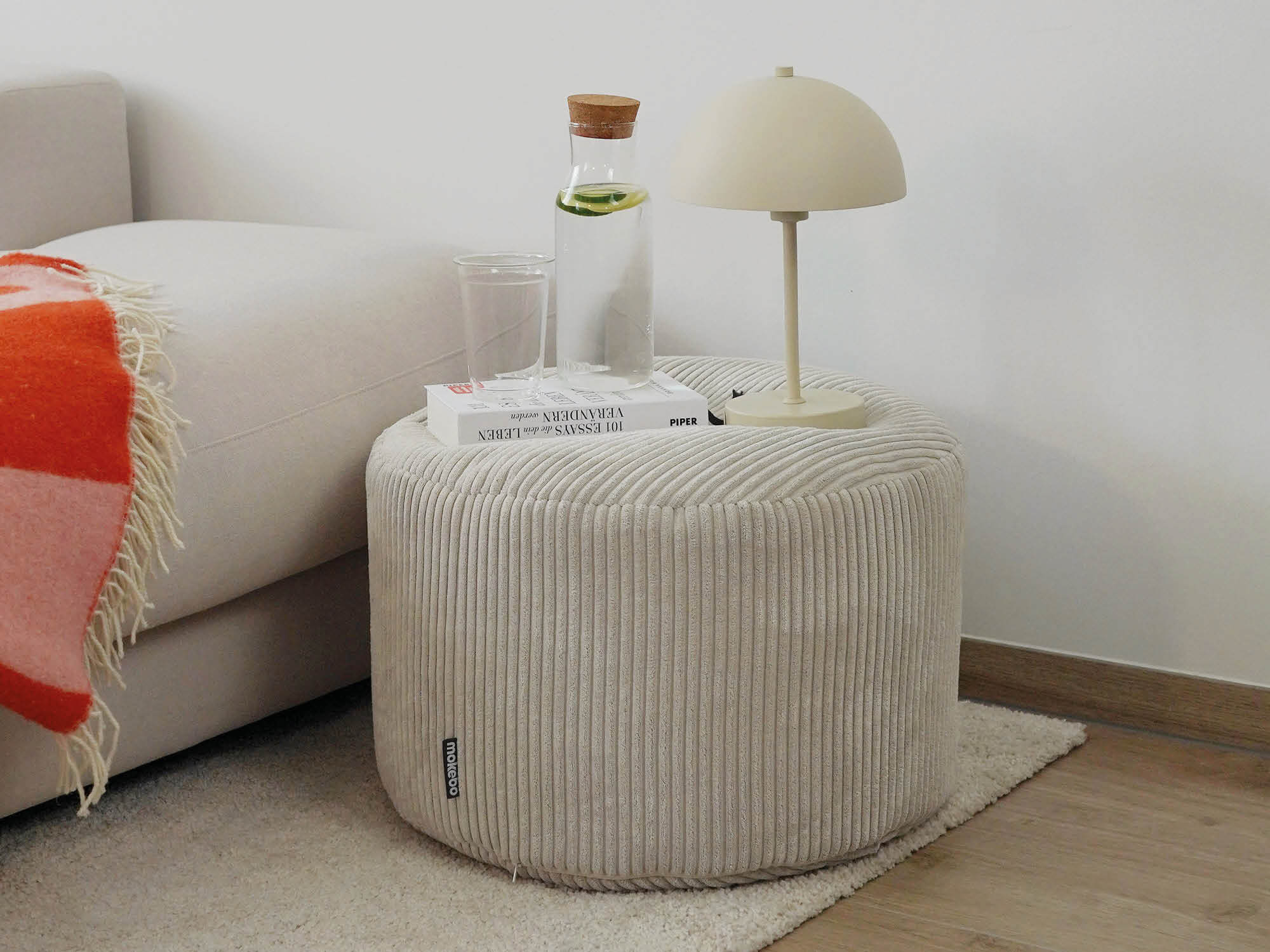 runder Pouf aus Cord in beige vor einem beigen Sofa. Auf dem Pouf ist eine weiße Kabellose Tischlampe, eine Wasserkaraffe mit Glas und ein Buch.