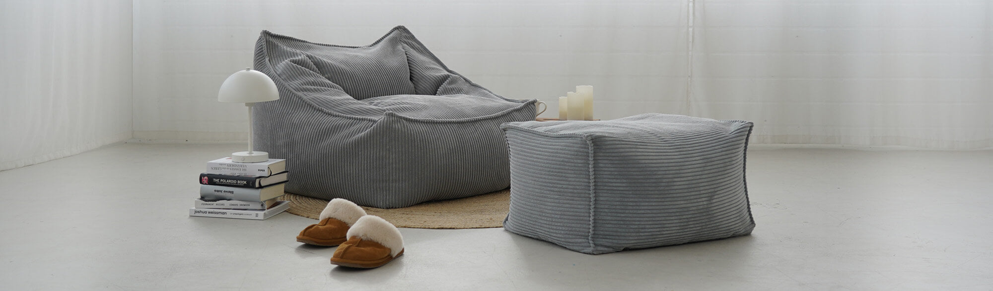 Sitzsack aus Cord in Hellgrau 'Der Lümmel' mit Pouf auf runden braunen Teppich, Bücherstapel mit Lampe daneben und braune Hausschuhe davor
