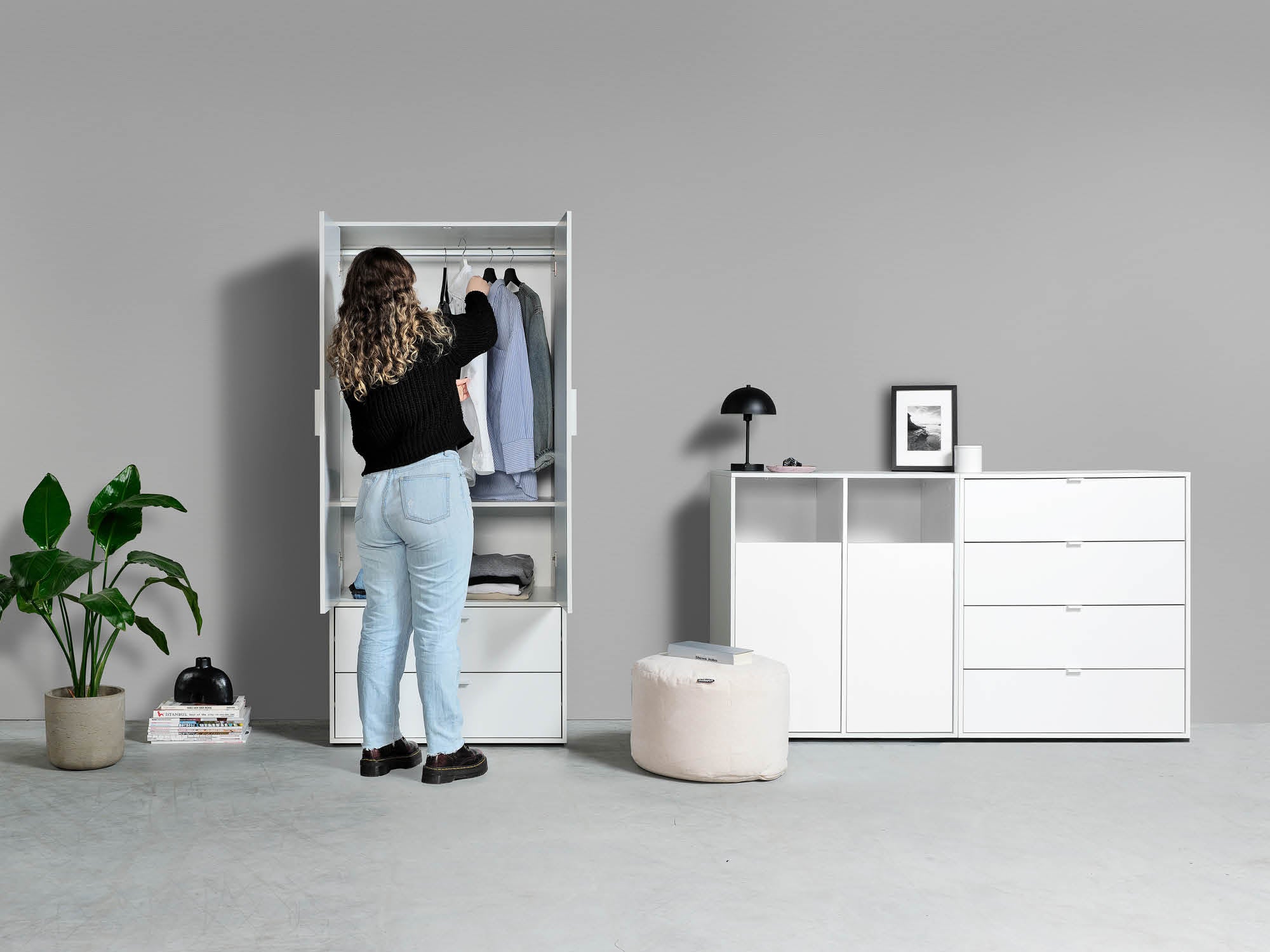 Geöffneter Kleiderschrank mit Kleiderstange und zwei Fächer unten. Frau steht vor dem Kleiderschrank und nimmt ein weißes Hemd heraus. weißer Wäscheschrank und weiße Kommode mit vier Fächern daneben.
