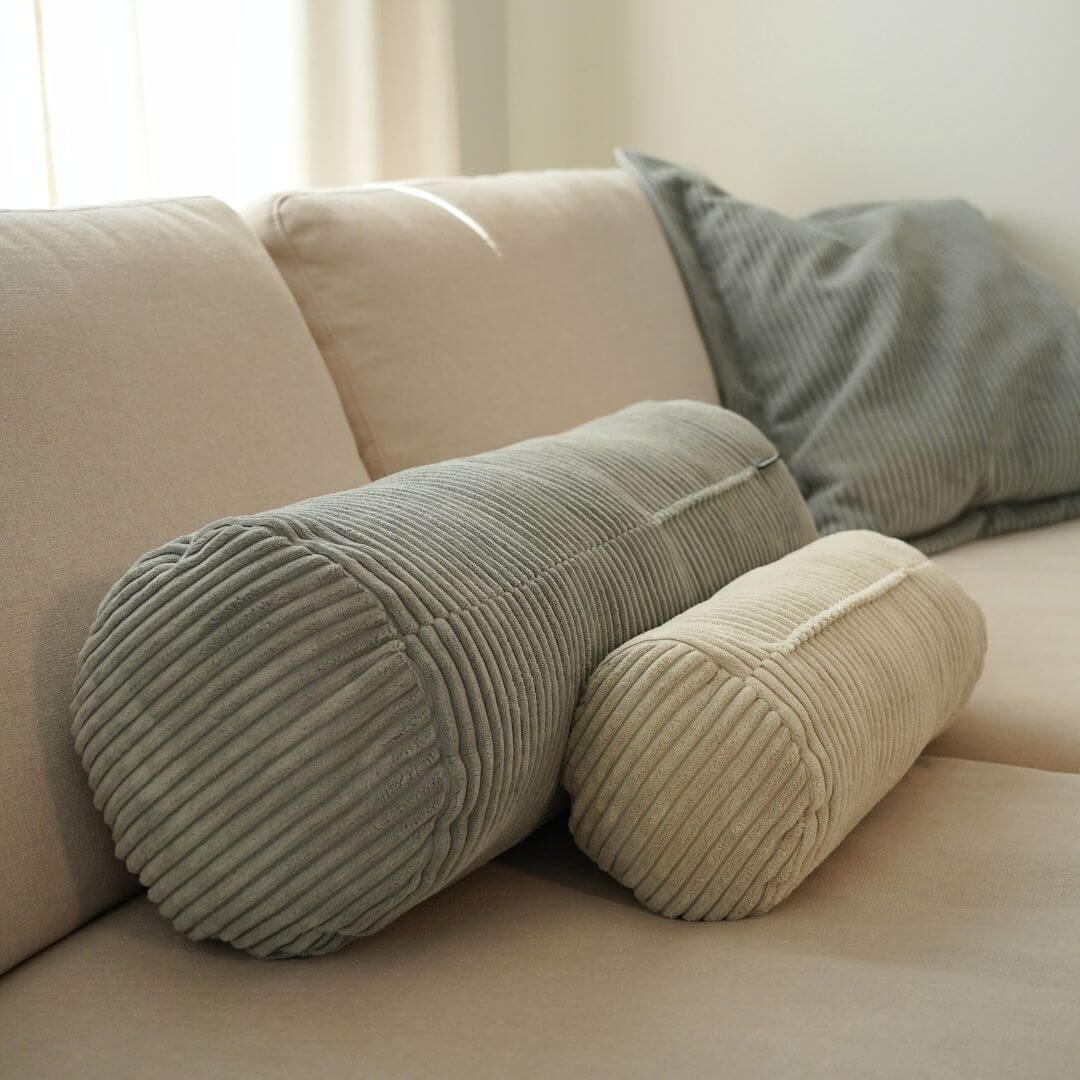 große graue und kleine beige  Nackenrolle aus Cord  hintereinander auf einem beigen Sofa von der Seite