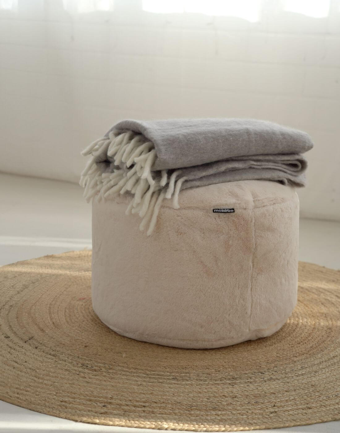 runder Pouf mit weichem Kunstfell in creme mit grauer Decke darauf.
