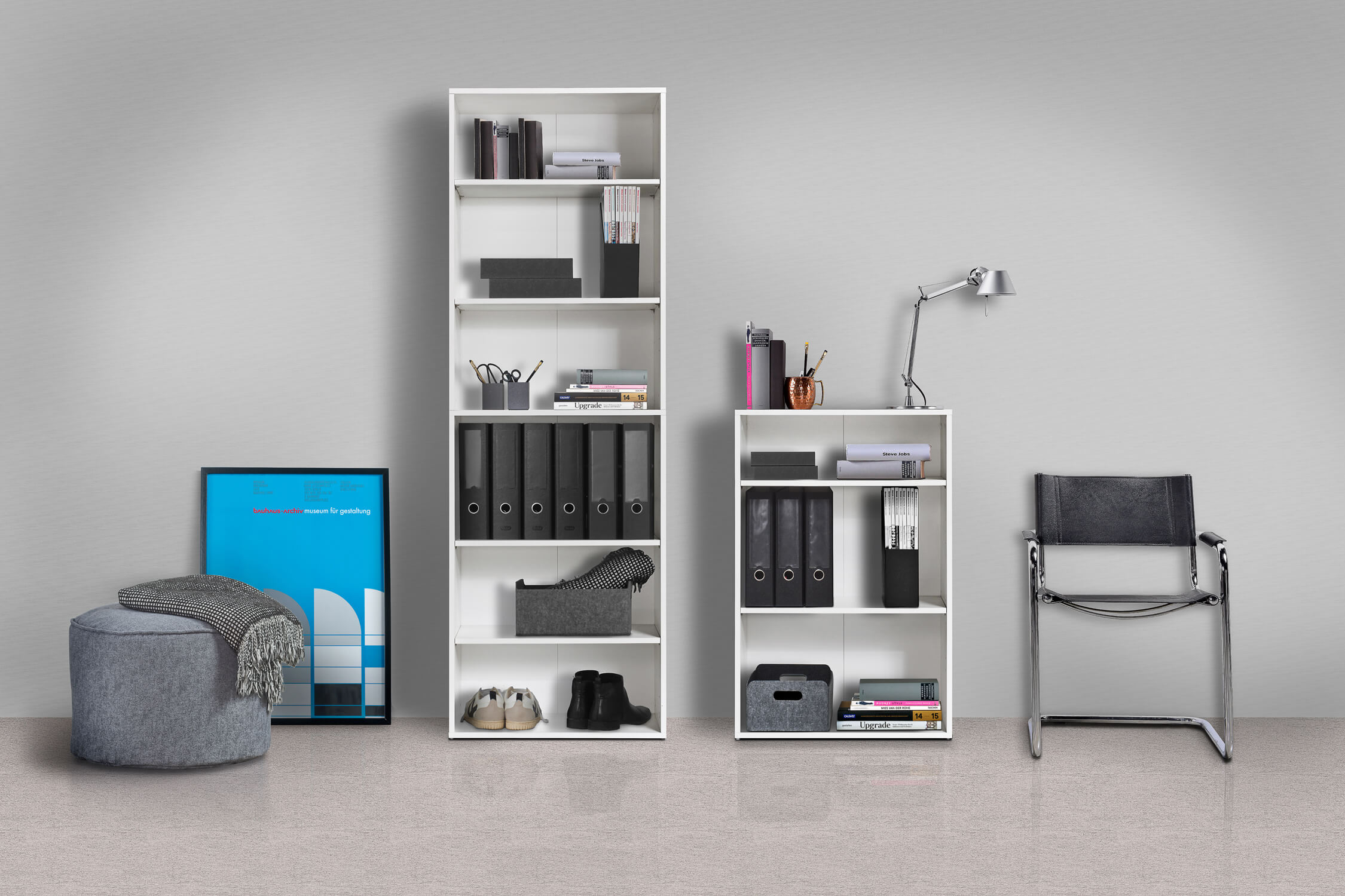 Ein hohes und ein niedriges Regal nebeneinander an einer Wand. Beide sind weiß und offen mit Akten, Büchern und Kisten drinnen.